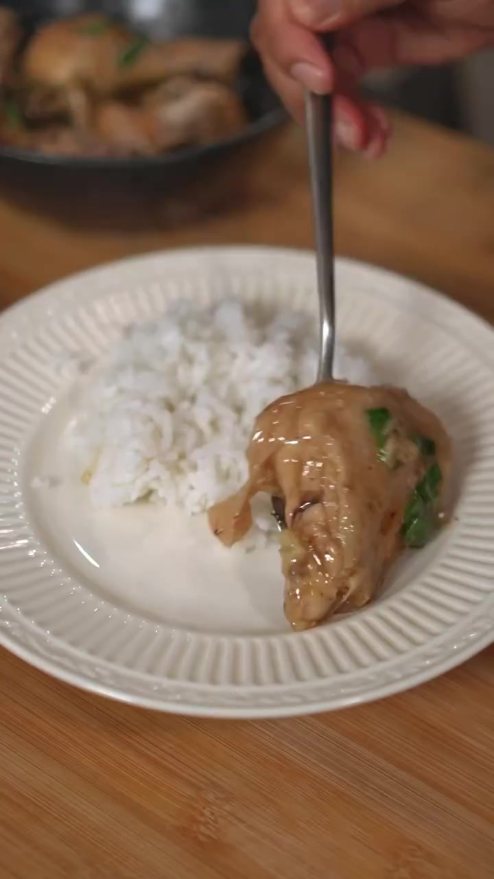 Sepotong ayam kukus berkuah gurih di sebelah nasi putih pada piring putih di atas meja kayu