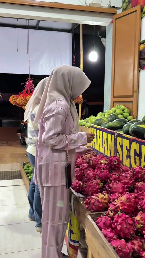 Tampak samping wanita berhijab memakai set piyama satin lilac bergaris di depan kios buah naga pasar malam