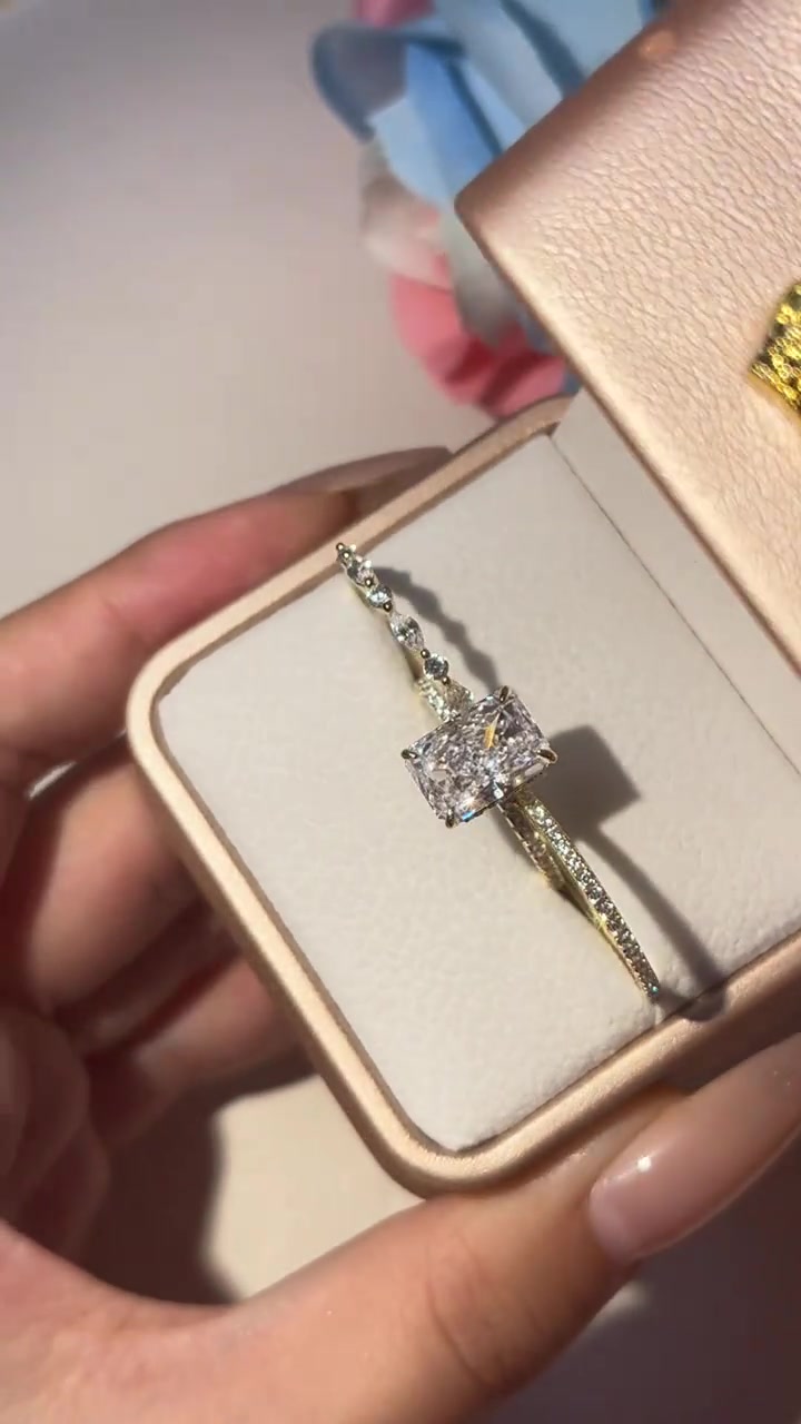 Cincin moissanite potongan radiant berangka gold di dalam kotak krem yang dipegang tangan