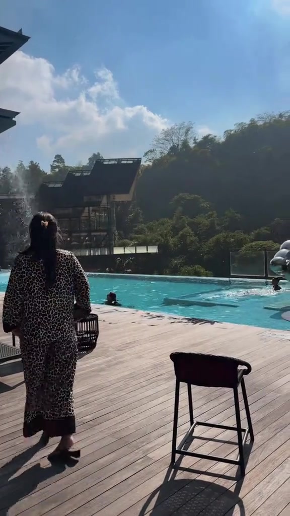 Tampak belakang wanita memakai set piyama satin motif leopard membawa tas anyaman di deck kolam renang hotel