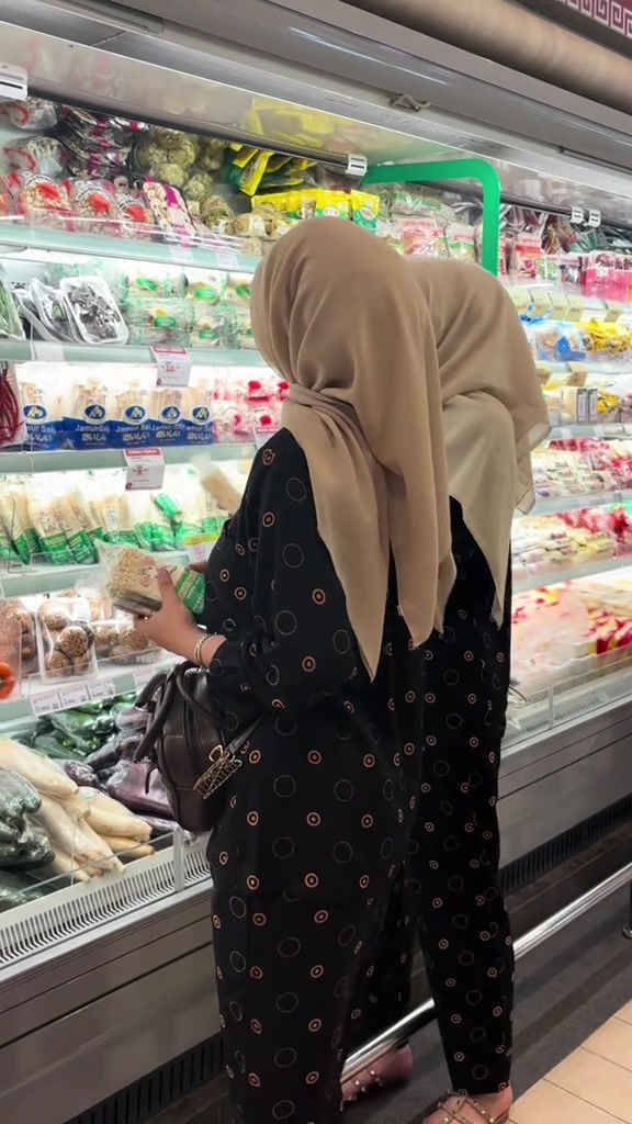 Dua wanita berhijab memakai set piyama hitam bermotif kecil, terlihat dari belakang saat memilih barang di depan chiller supermarket