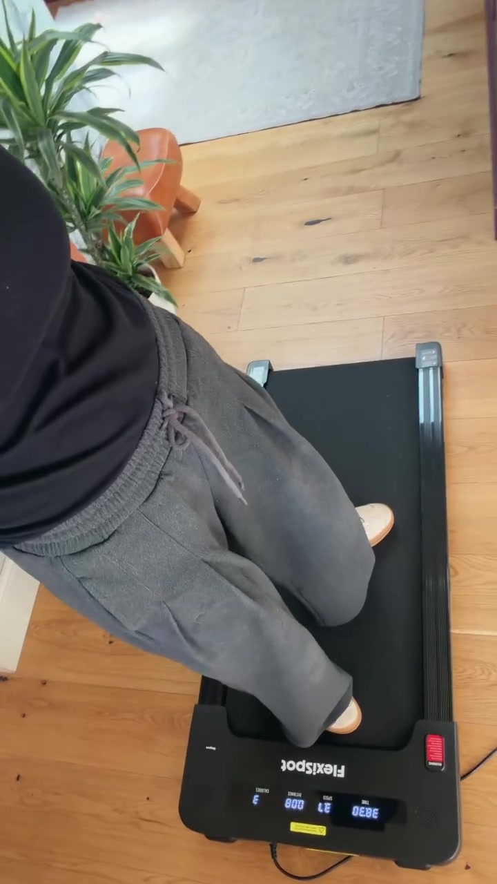 Tampak atas orang bercelana panjang abu-abu berjalan di atas walking pad dengan layar LED menyala