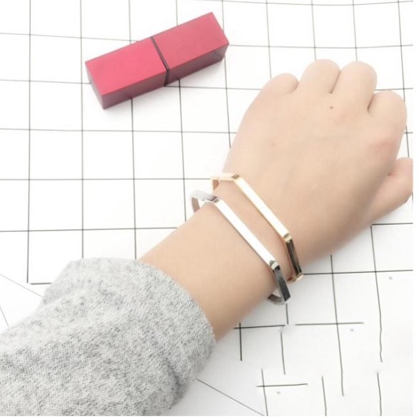 Dua gelang bangle tipis warna gold dan silver dipakai bersamaan di pergelangan tangan di atas latar kertas kotak