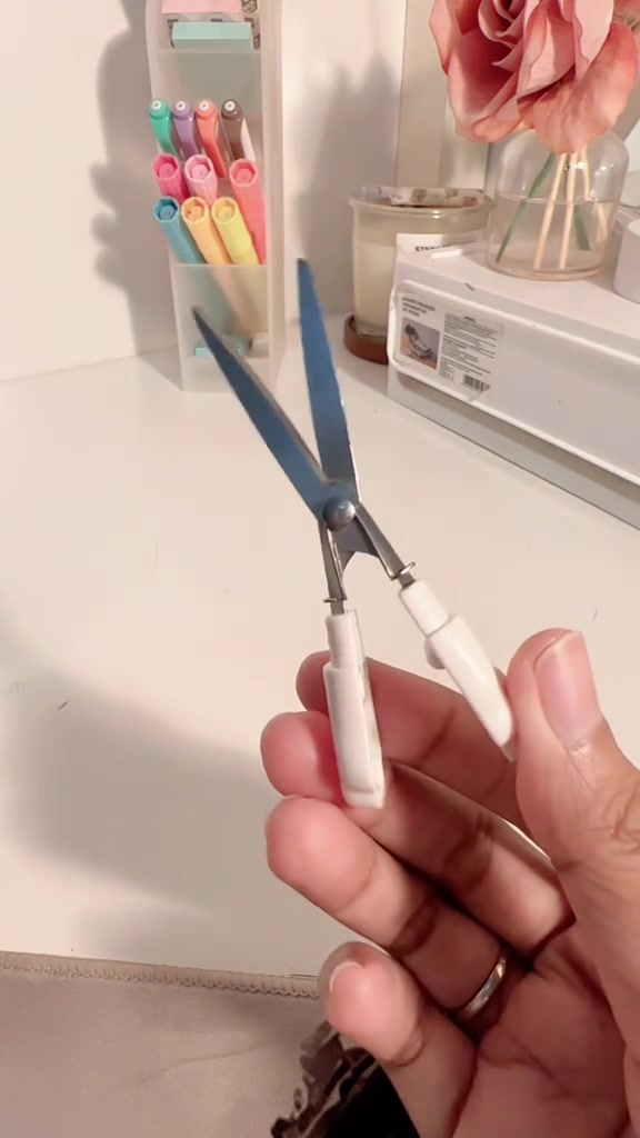Tangan memegang gunting lipat kecil yang terbuka di atas meja belajar