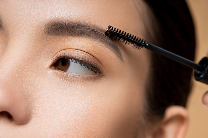 Close-up bulu alis gelap sedang disisir dengan aplikator spoolie browcara