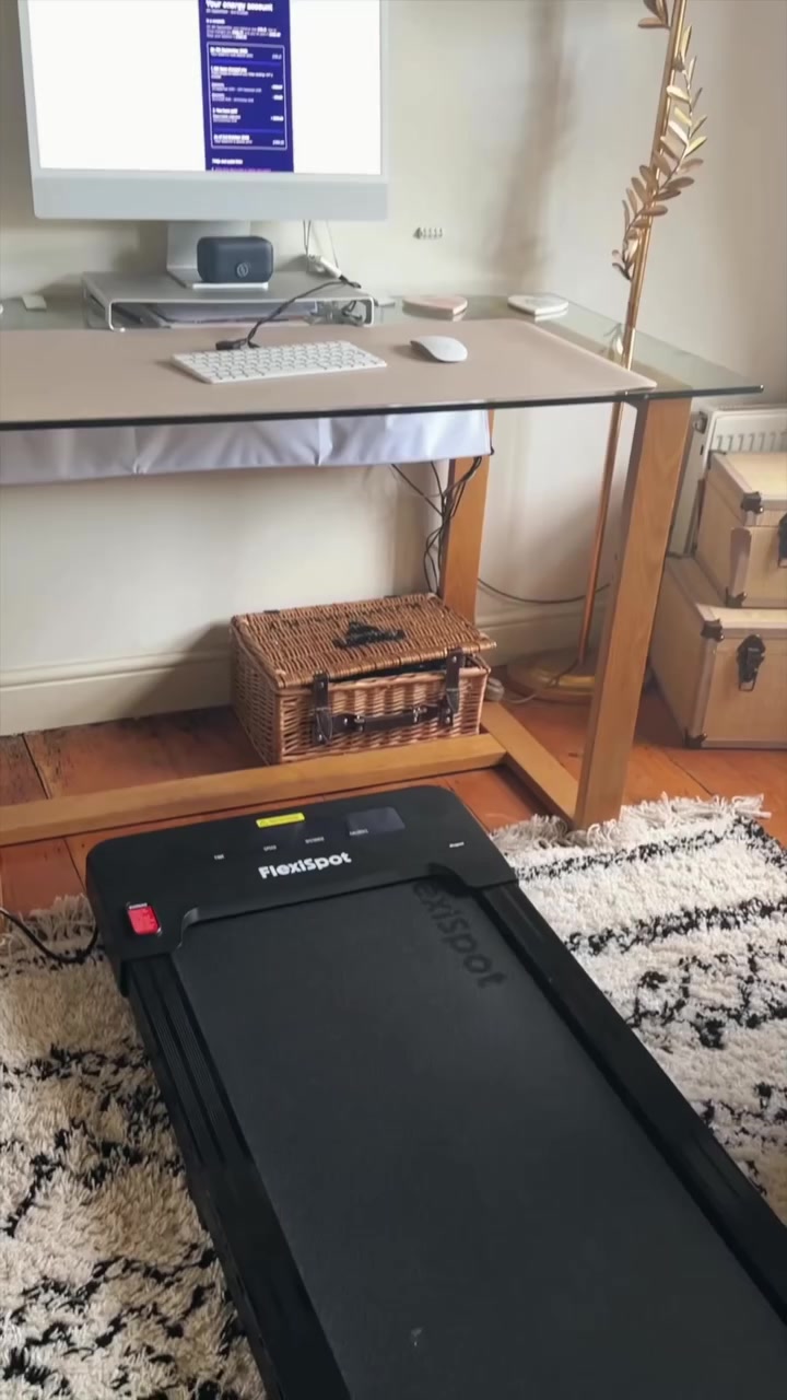 Walking pad hitam tersimpan rapi di bawah meja kerja kaca di ruang kerja rumah