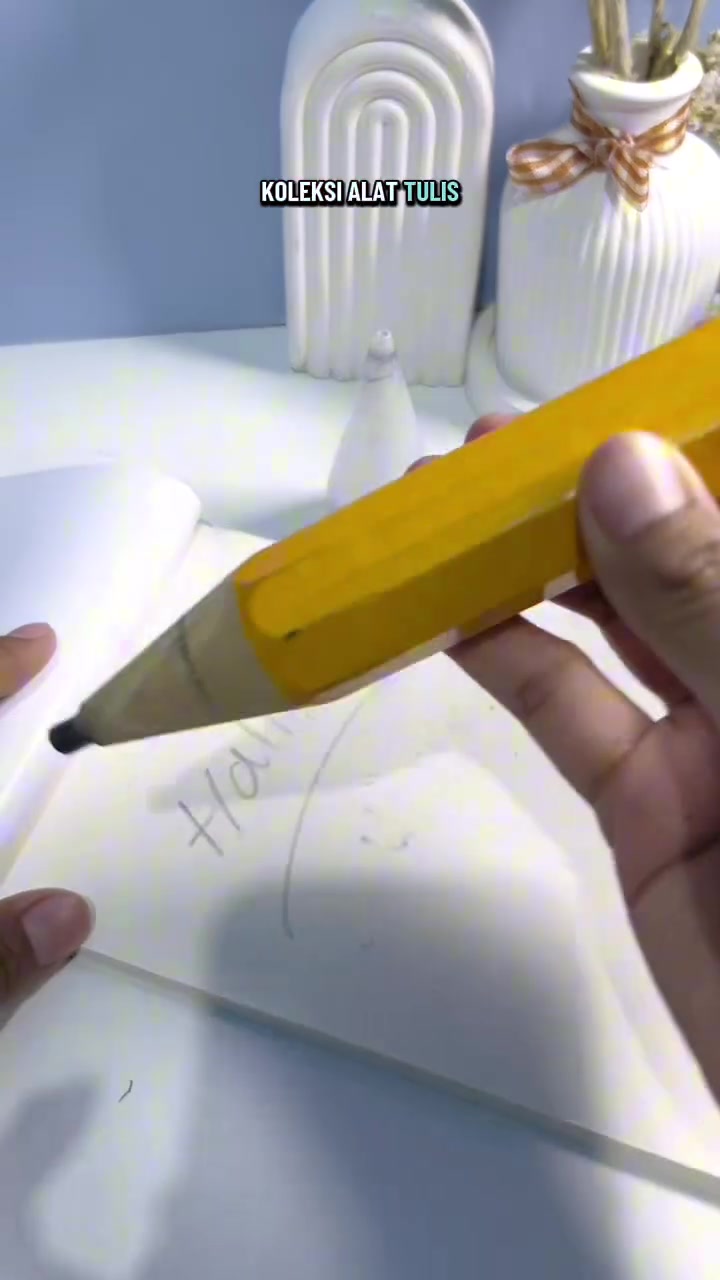 Tangan memegang pensil jumbo kuning dekat halaman buku yang terbuka.