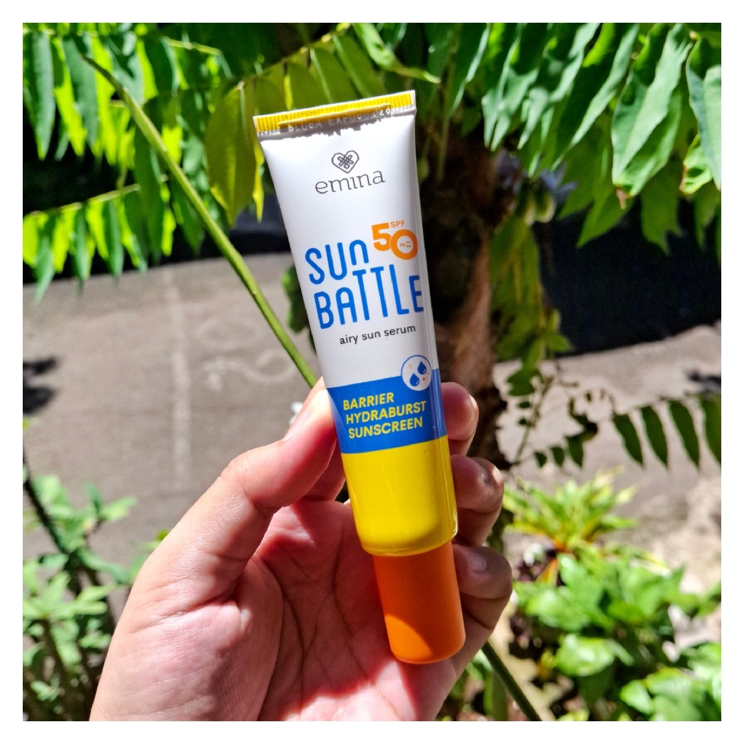 Tube Emina Sun Battle Barrier Hydraburst Sunscreen SPF 50 dipegang dengan latar tanaman hijau
