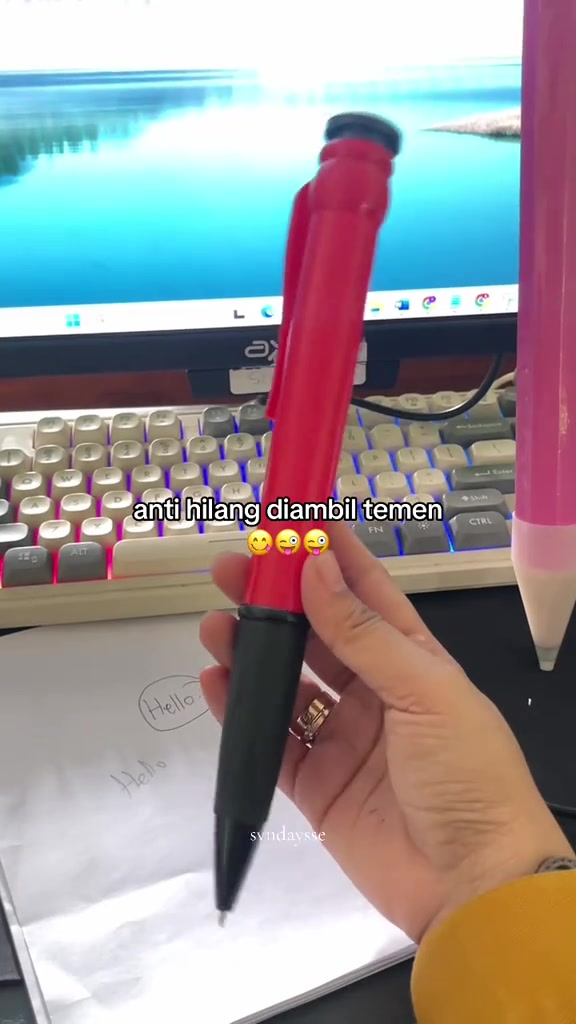Tangan memegang pulpen jumbo merah di depan keyboard, dengan pensil besar merah muda di sampingnya.