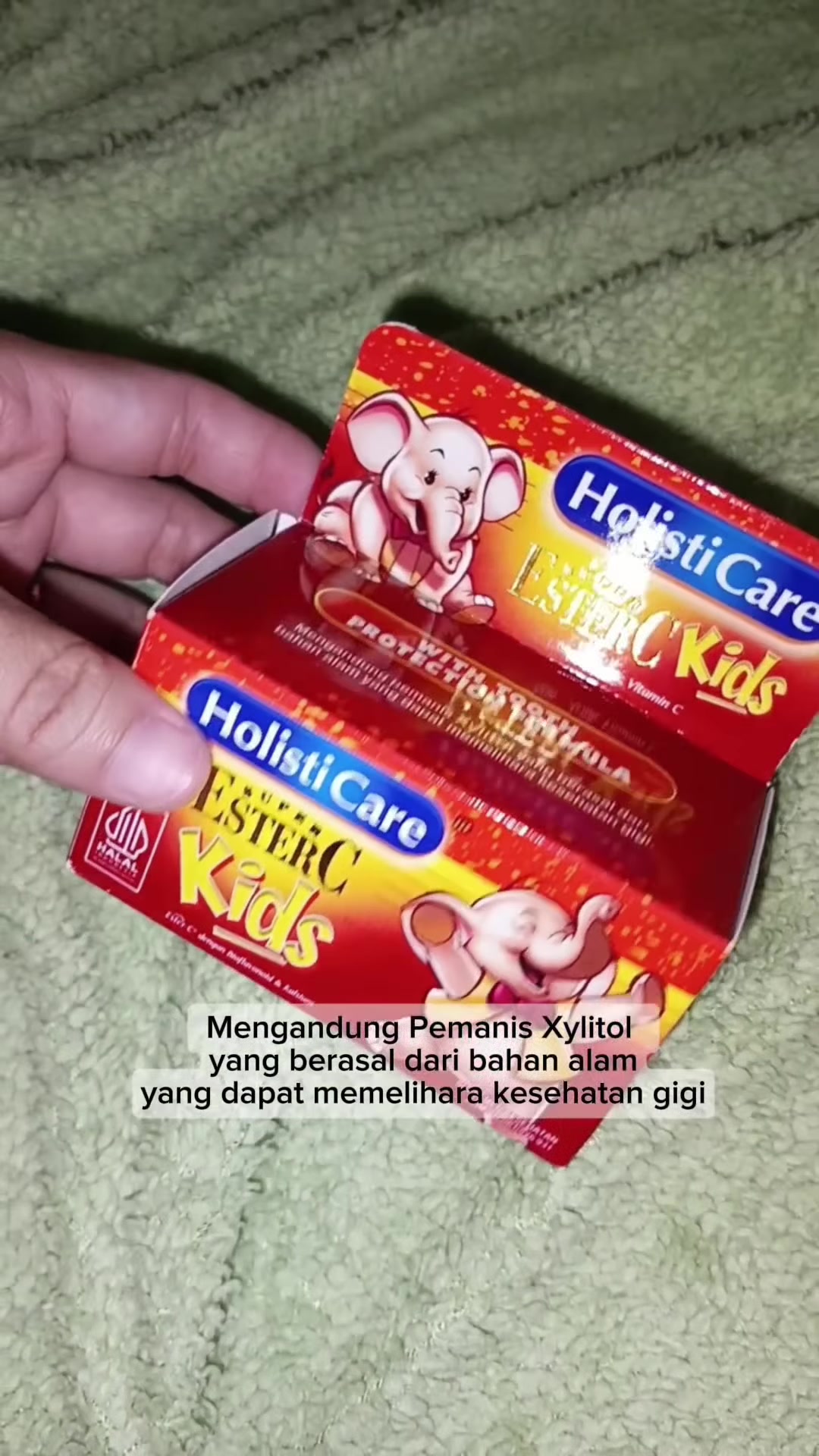 Tangan memegang kotak Holisticare Ester-C Kids berwarna merah dan kuning.