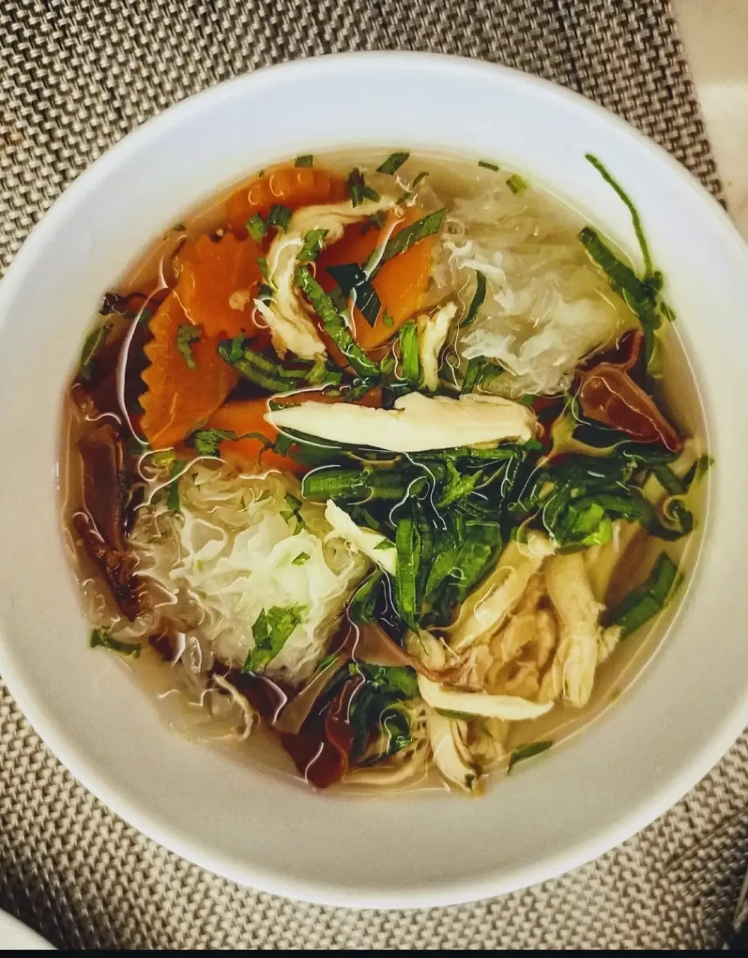 Semangkuk sup bening ayam dan sayuran dengan wortel serta daun hijau di atas alas tenun