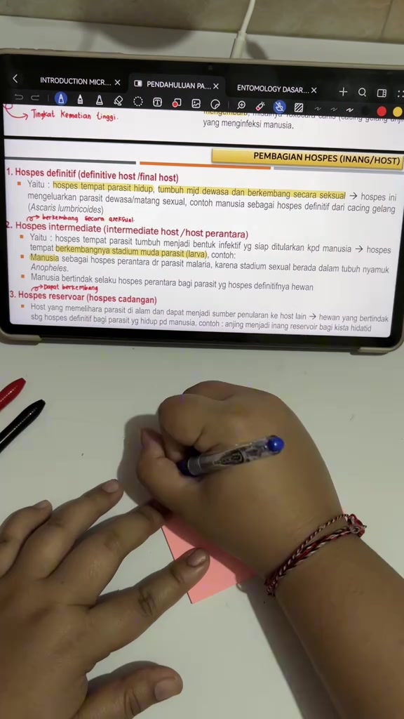 Sticky note merah muda sedang ditulis di meja belajar di bawah layar catatan digital