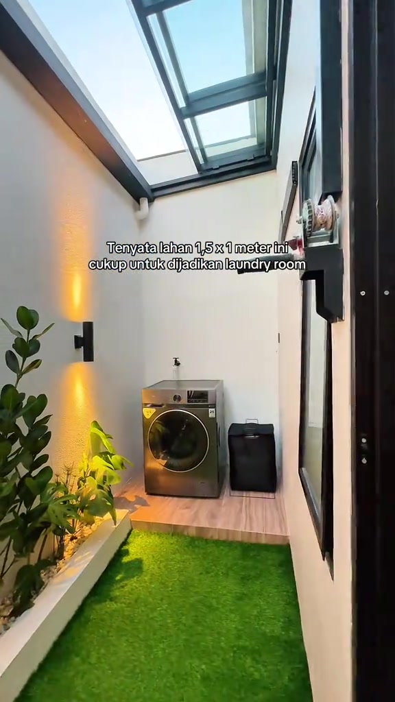 Sudut laundry mungil dengan mesin cuci, tanaman di sisi kiri, dan bukaan atap yang menerangi ruang