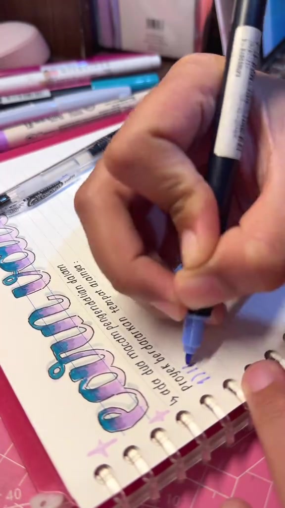Tangan menulis di buku catatan spiral dengan judul berwarna biru dan ungu