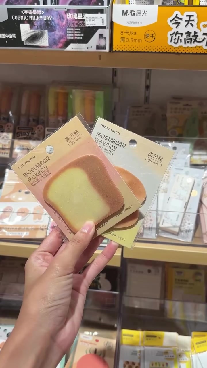 Tangan memegang beberapa memo pad warna pastel di depan rak alat tulis