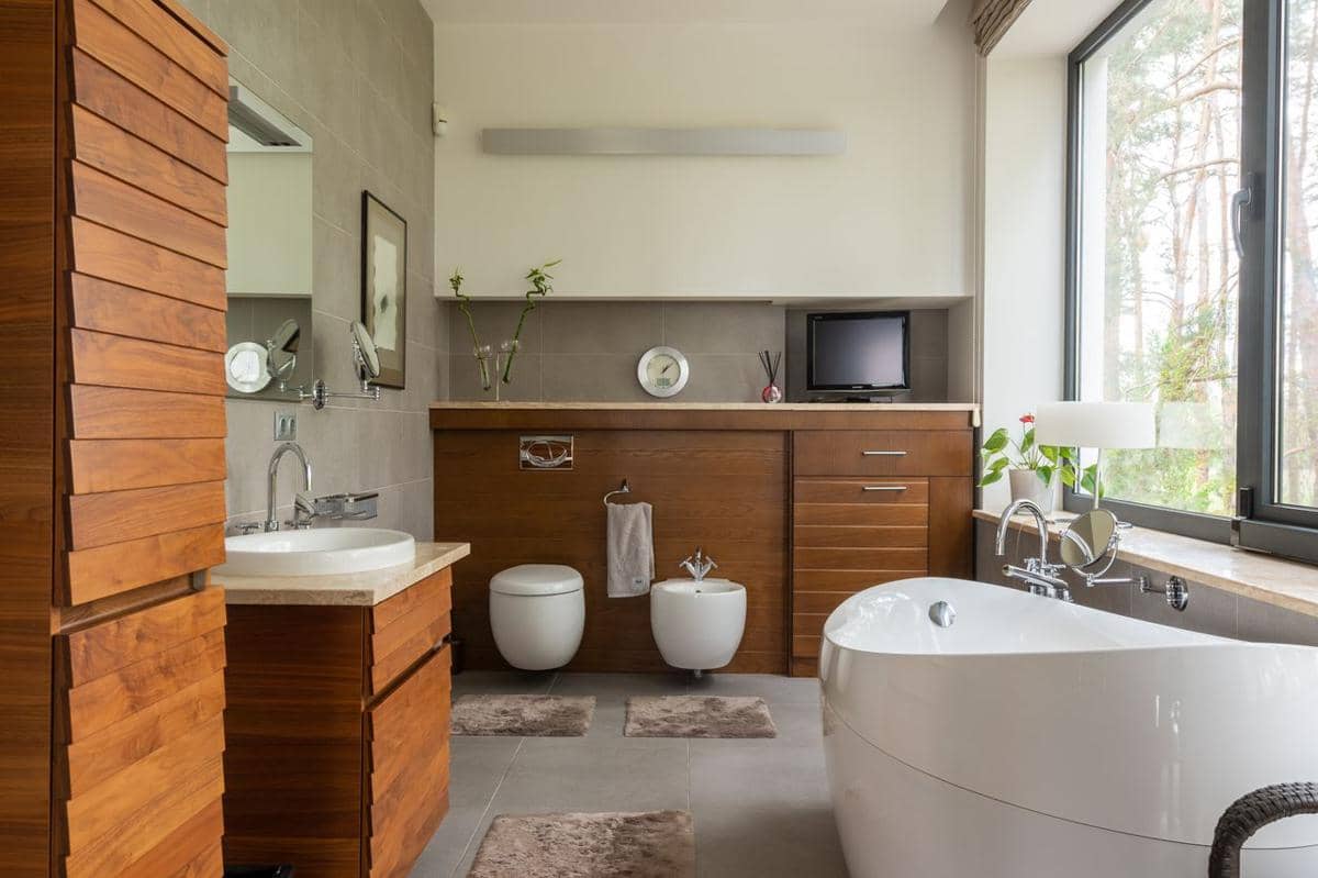 Kamar mandi luas dengan kabinet kayu, permukaan abu-abu, wastafel, dan cahaya alami dari jendela