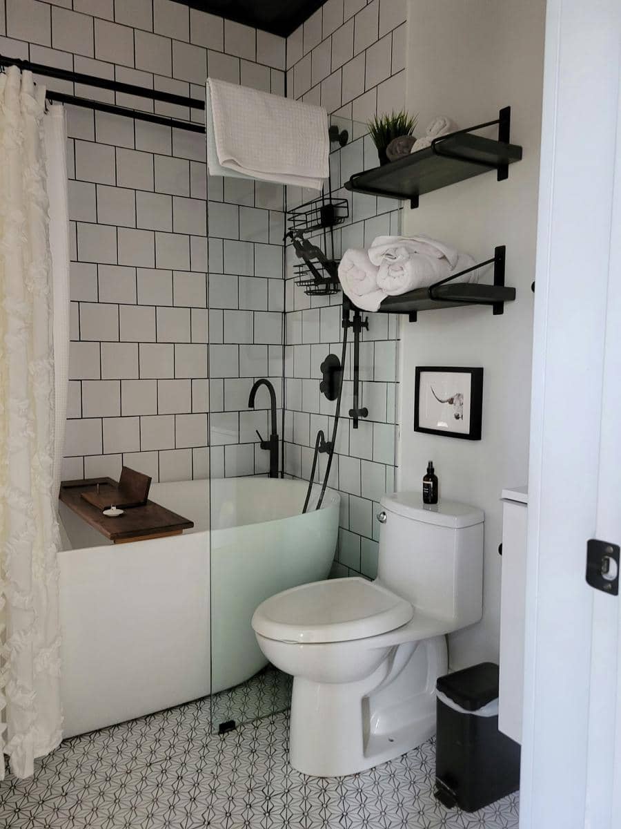 Kamar mandi bernuansa putih dengan rak hitam bertingkat di atas toilet dan area mandi