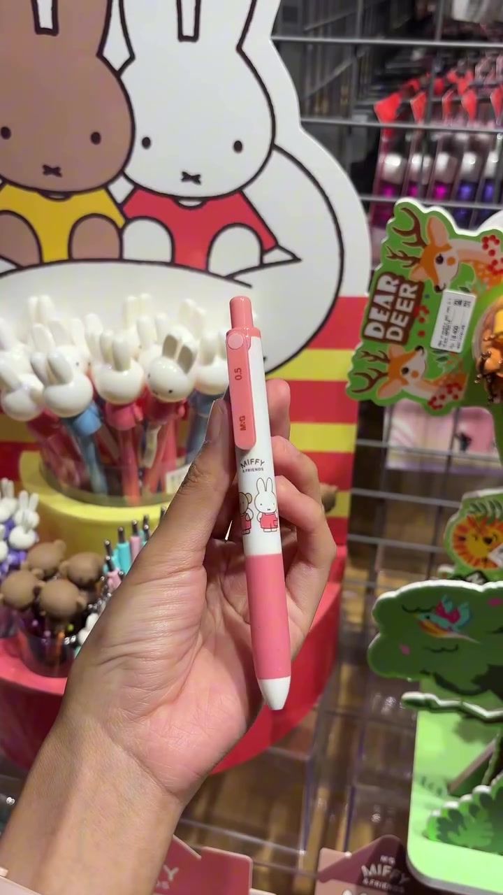 Tangan memegang pulpen gel Miffy warna merah muda di depan rak alat tulis