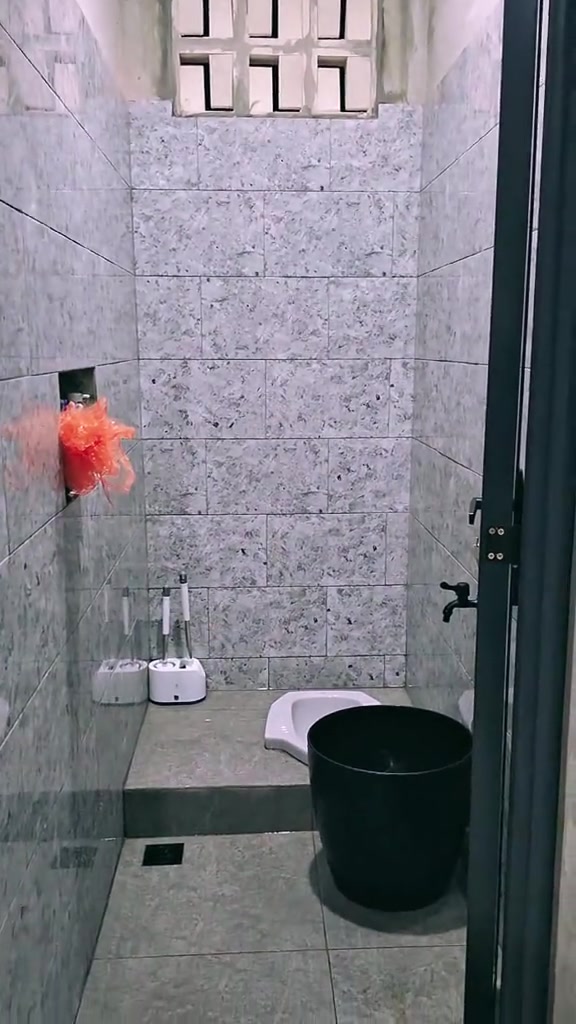 Kamar mandi kecil dengan ubin abu-abu, kloset jongkok, ventilasi, dan ember di sisi dinding
